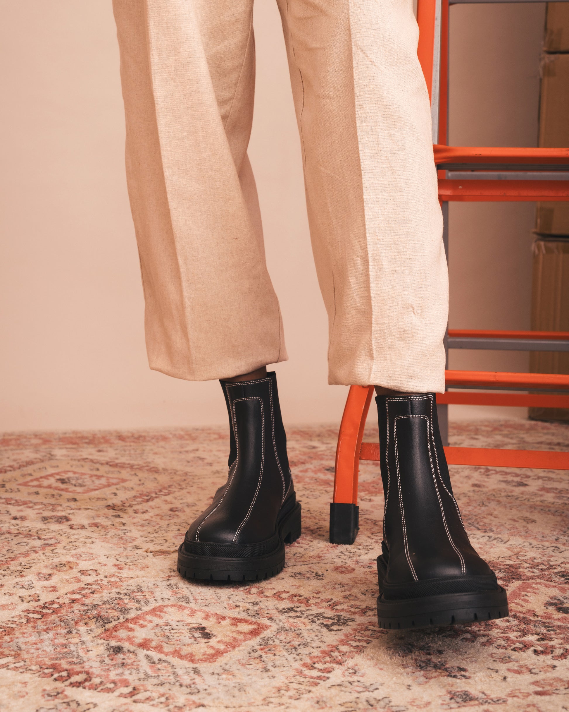 Person wearing black boots and beige pants on a patterned rug with an orange chair in the background.