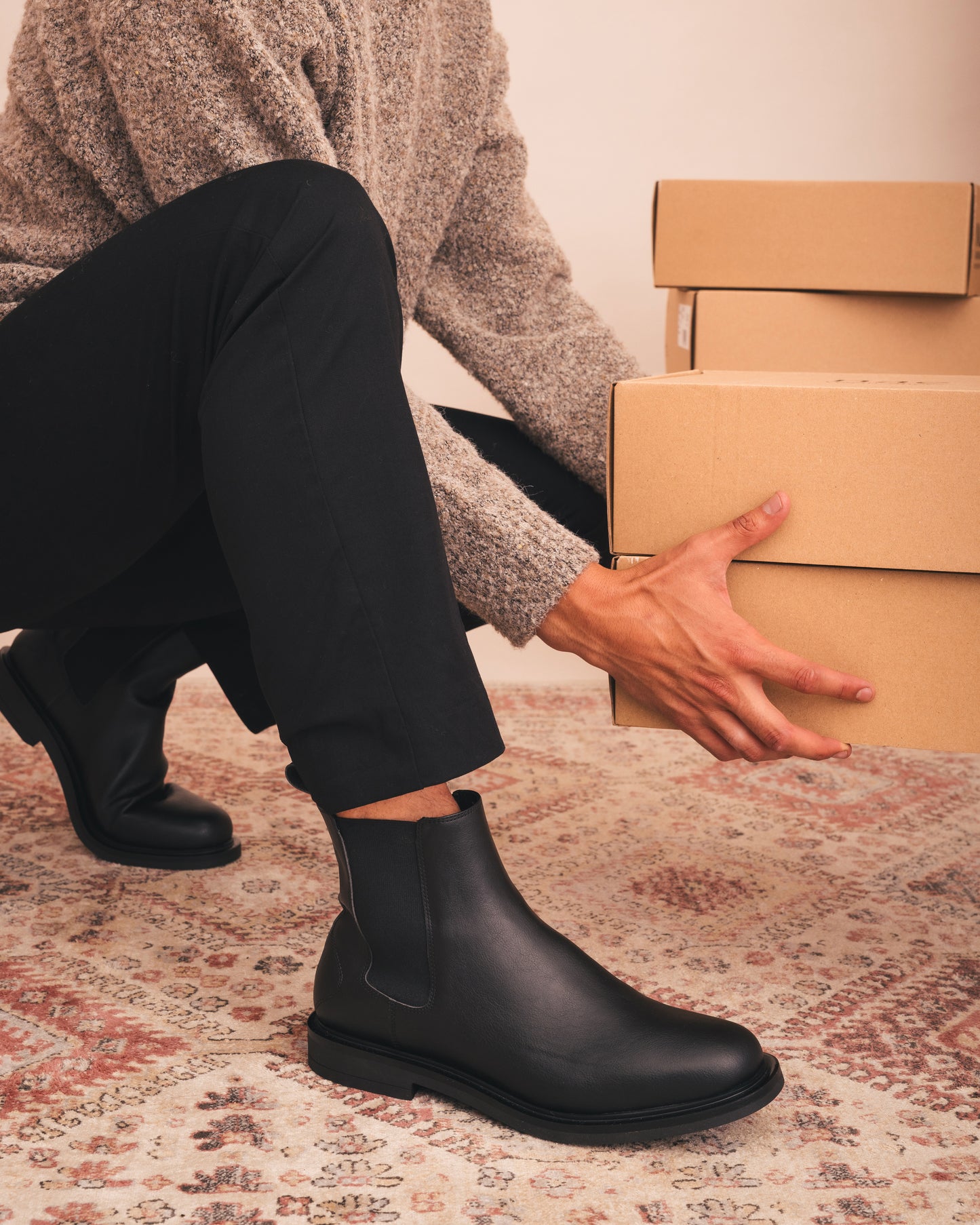 Person wearing black boots and holding cardboard boxes on a patterned rug.