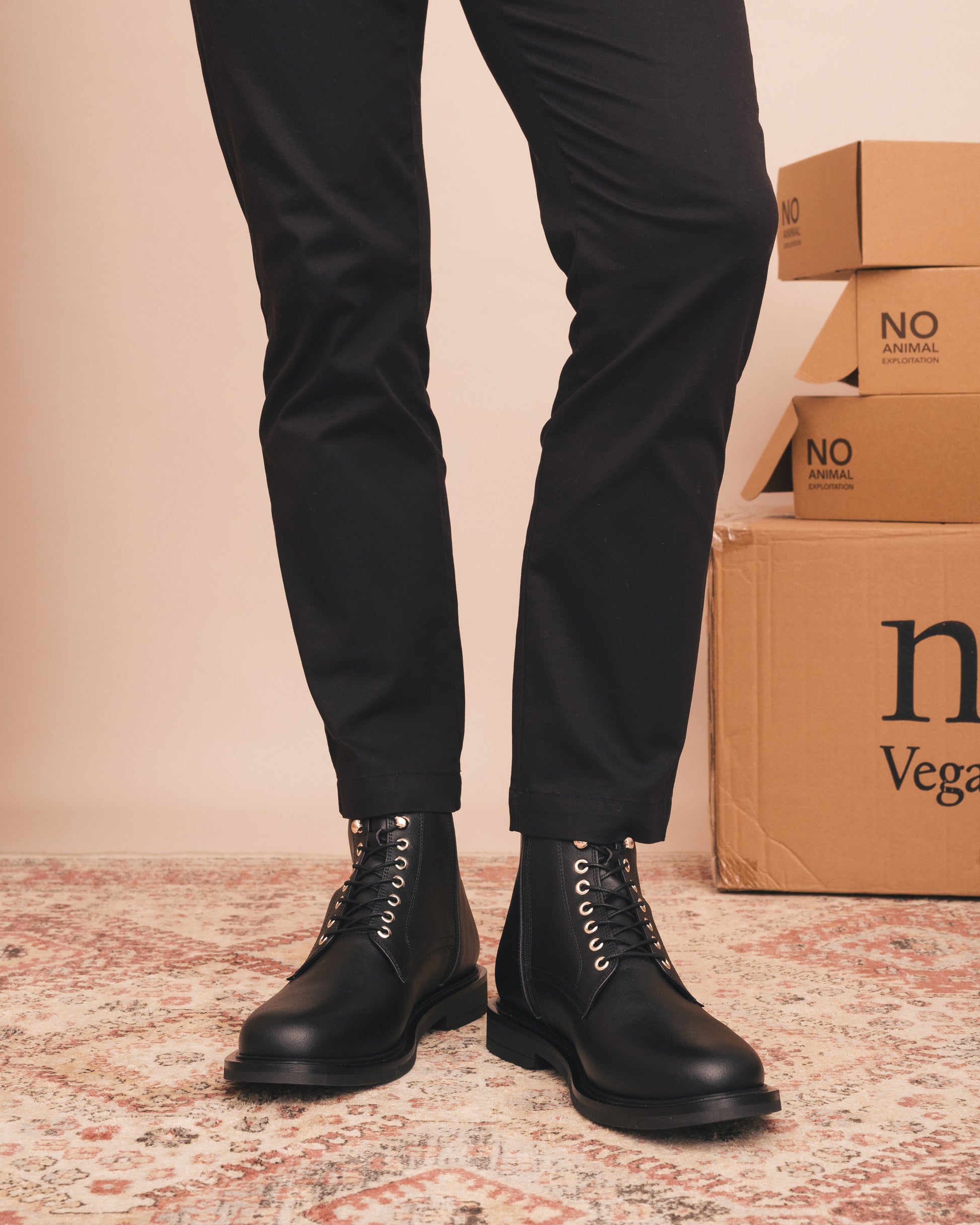Person wearing black boots and black pants standing next to cardboard boxes with 'No Animal' text on a patterned floor.