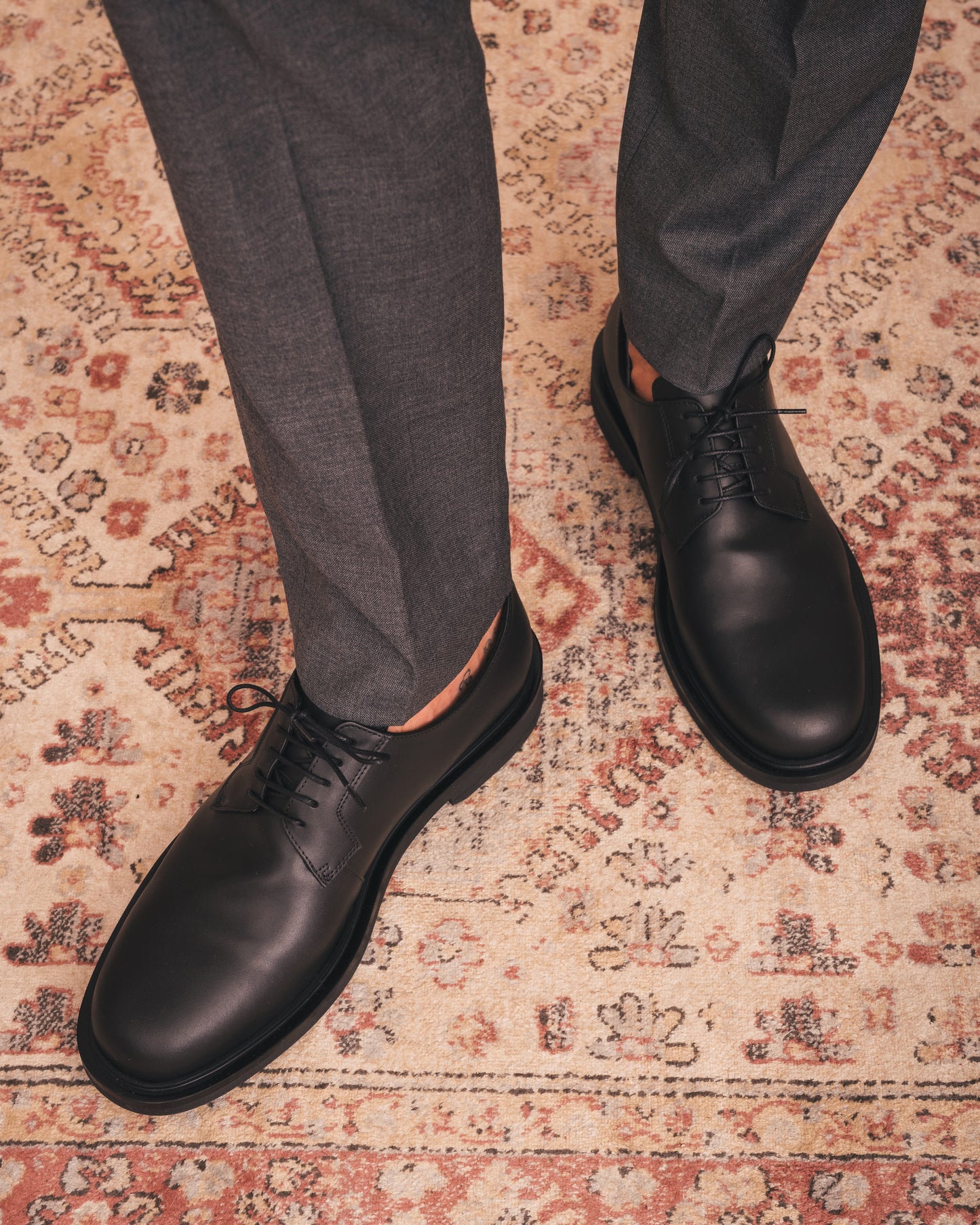 Black leather shoes worn with gray pants on a patterned rug