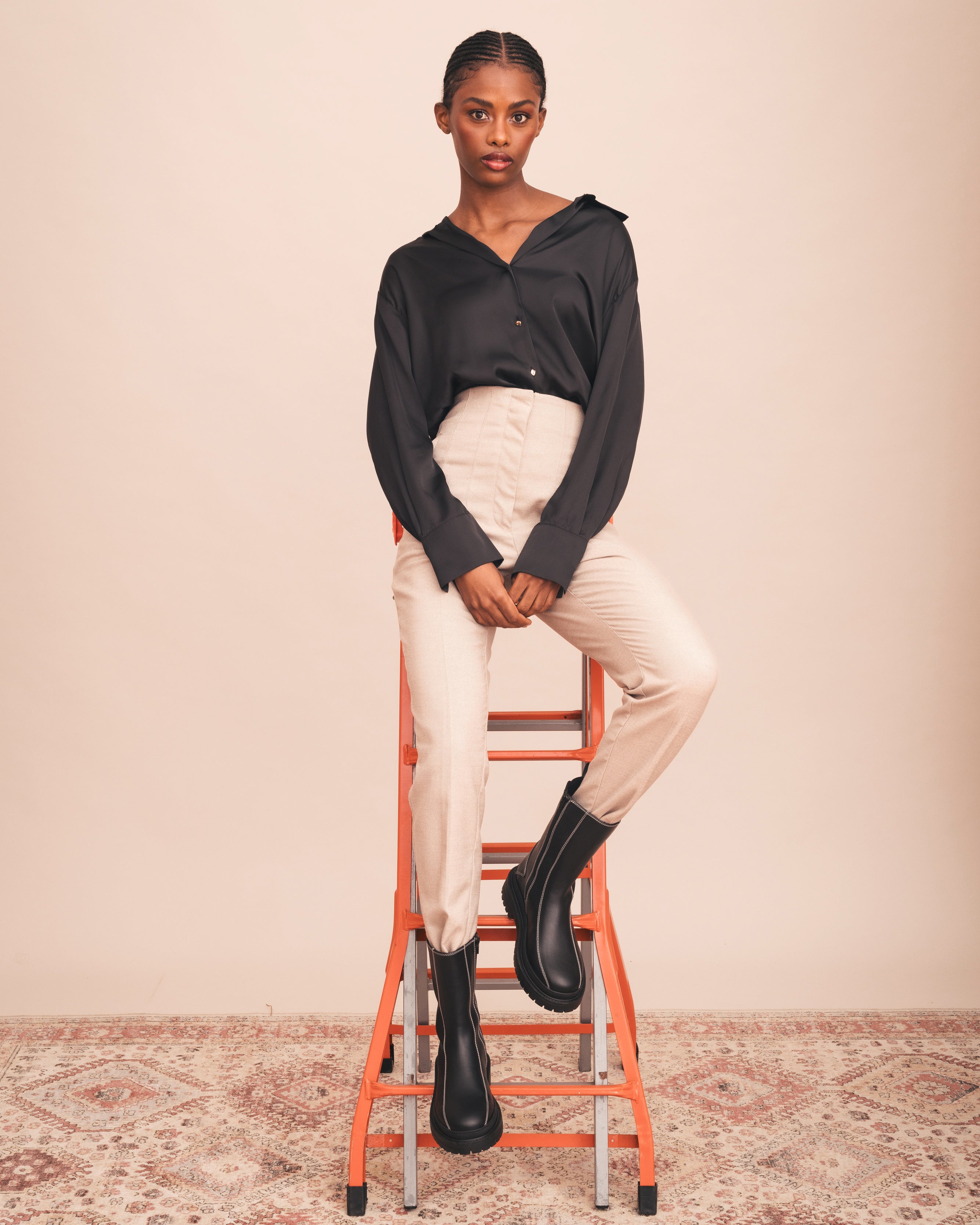 Woman sitting on an orange ladder wearing a black top and beige pants with black shoes against a plain background.