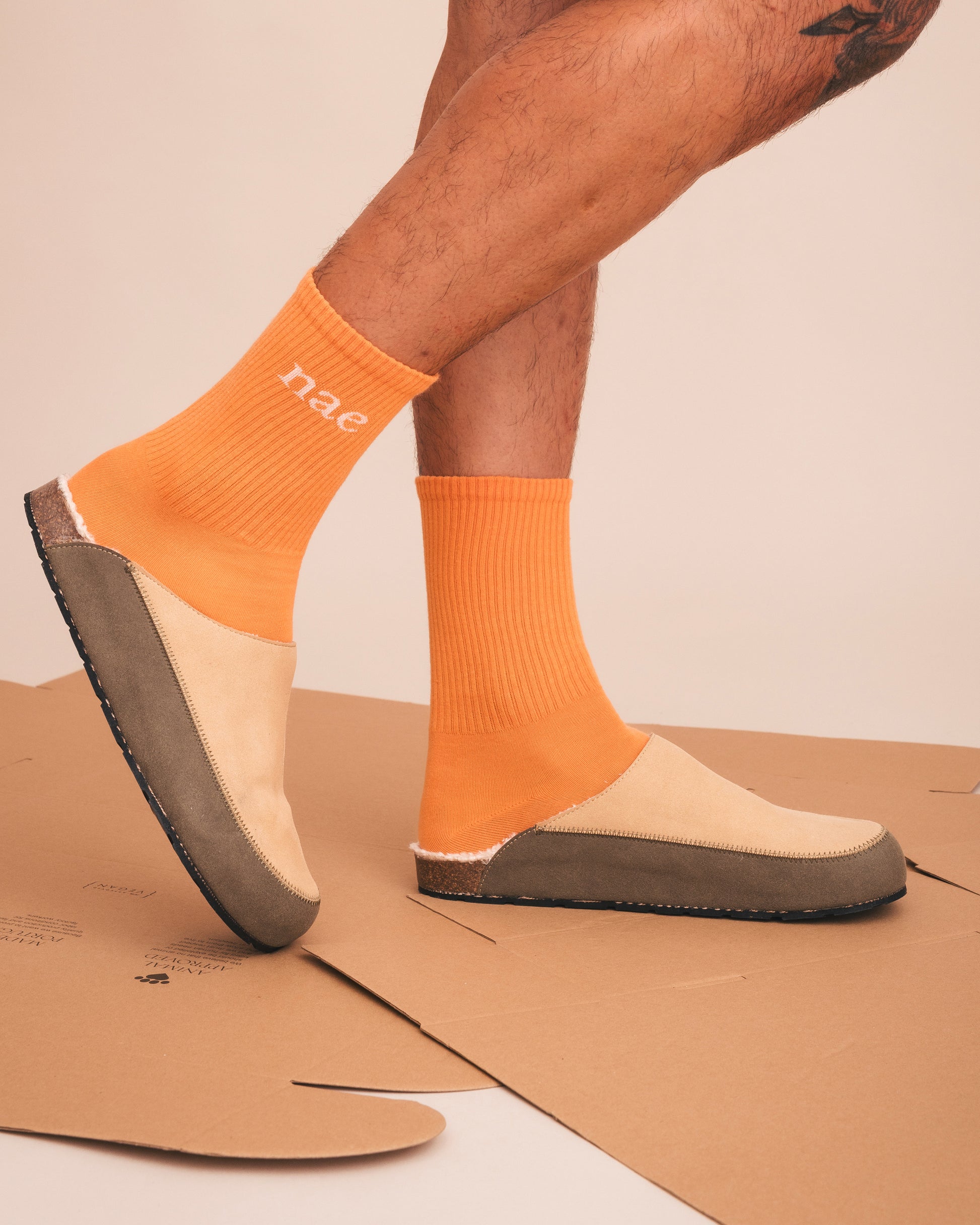 Person wearing orange socks with 'nae' branding and beige shoes on a cardboard surface.