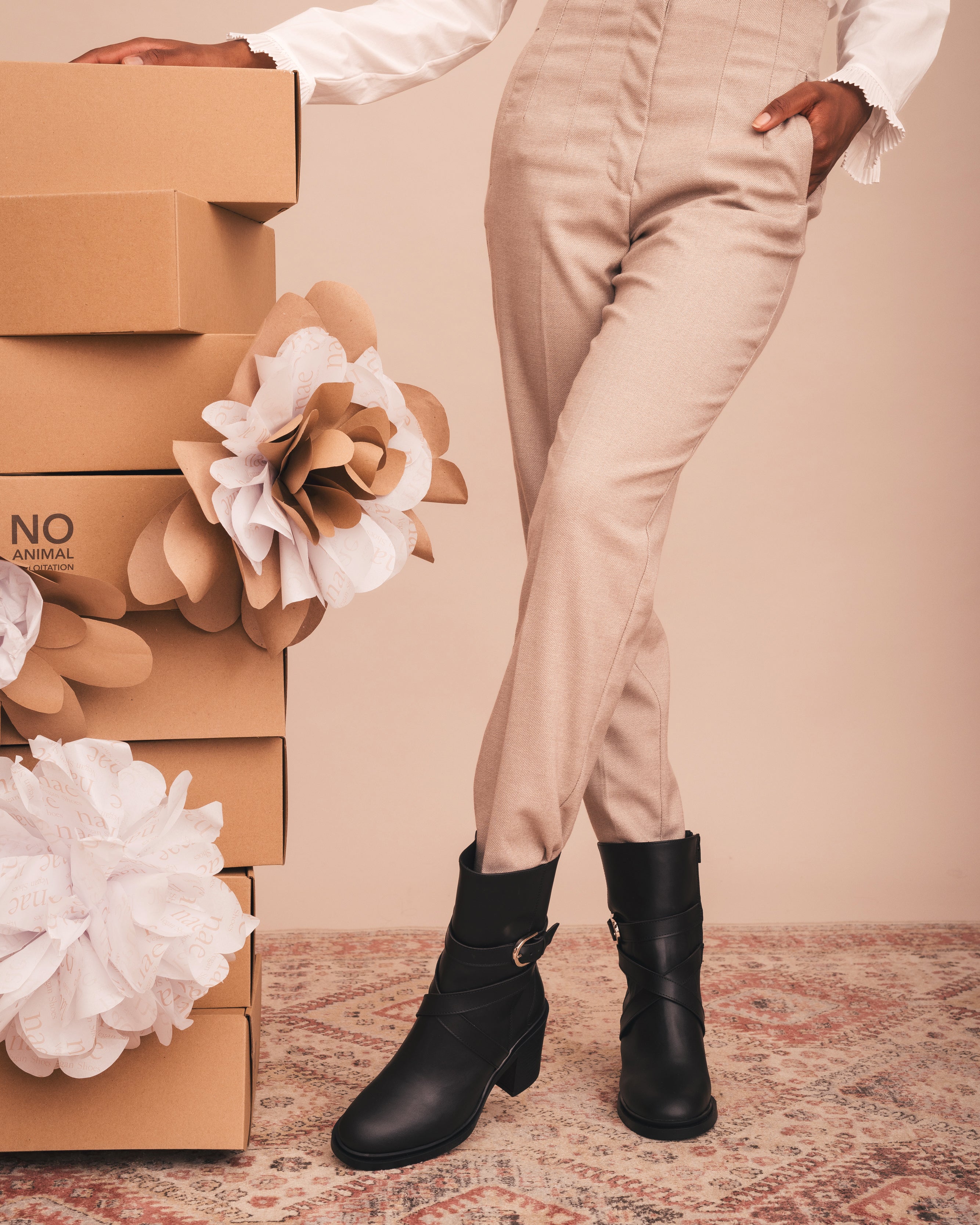 Person wearing black boots with stacked boxes and decorative flowers on a beige background