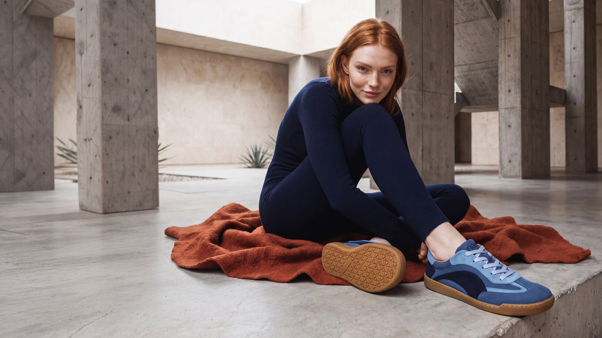 A woman with red hair sits on a rust-colored blanket on a concrete floor, wearing a dark navy long-sleeve top, black leggings, and blue suede sneakers with brown soles. She is posed in a minimalist concrete structure.