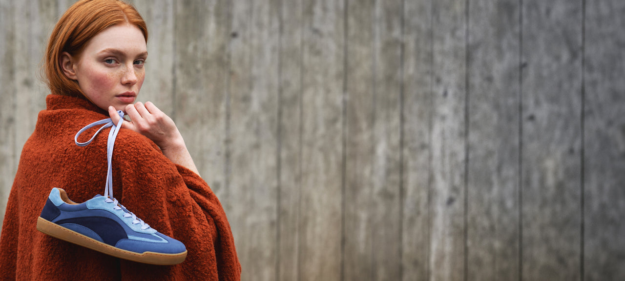 A red-haired woman wrapped in a rust-orange blanket stands against a weathered gray wooden wall, holding a blue suede sneaker with white laces and a gum sole.