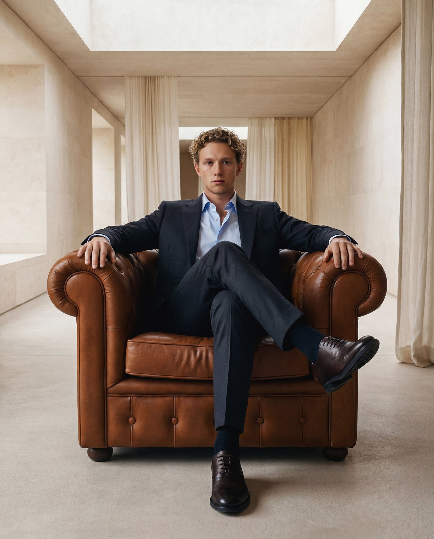 Man in a suit sitting on a brown leather armchair in a bright room with high ceilings.