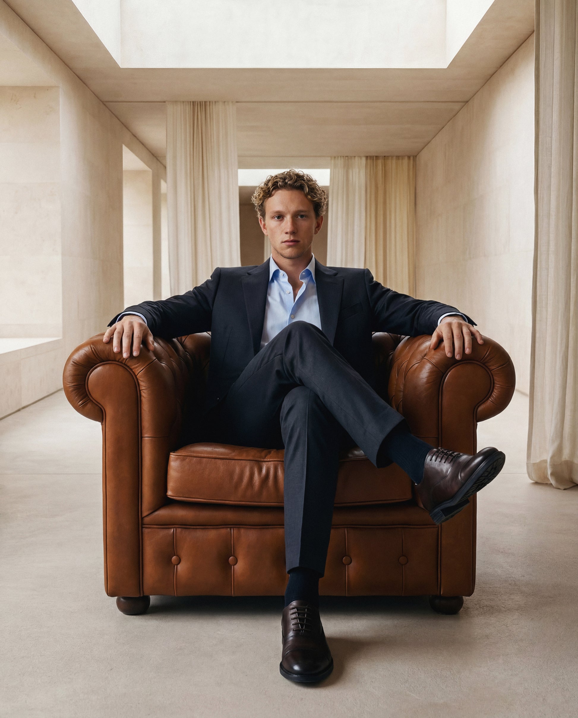 Man in a suit sitting on a brown leather armchair in a bright room with high ceilings.
