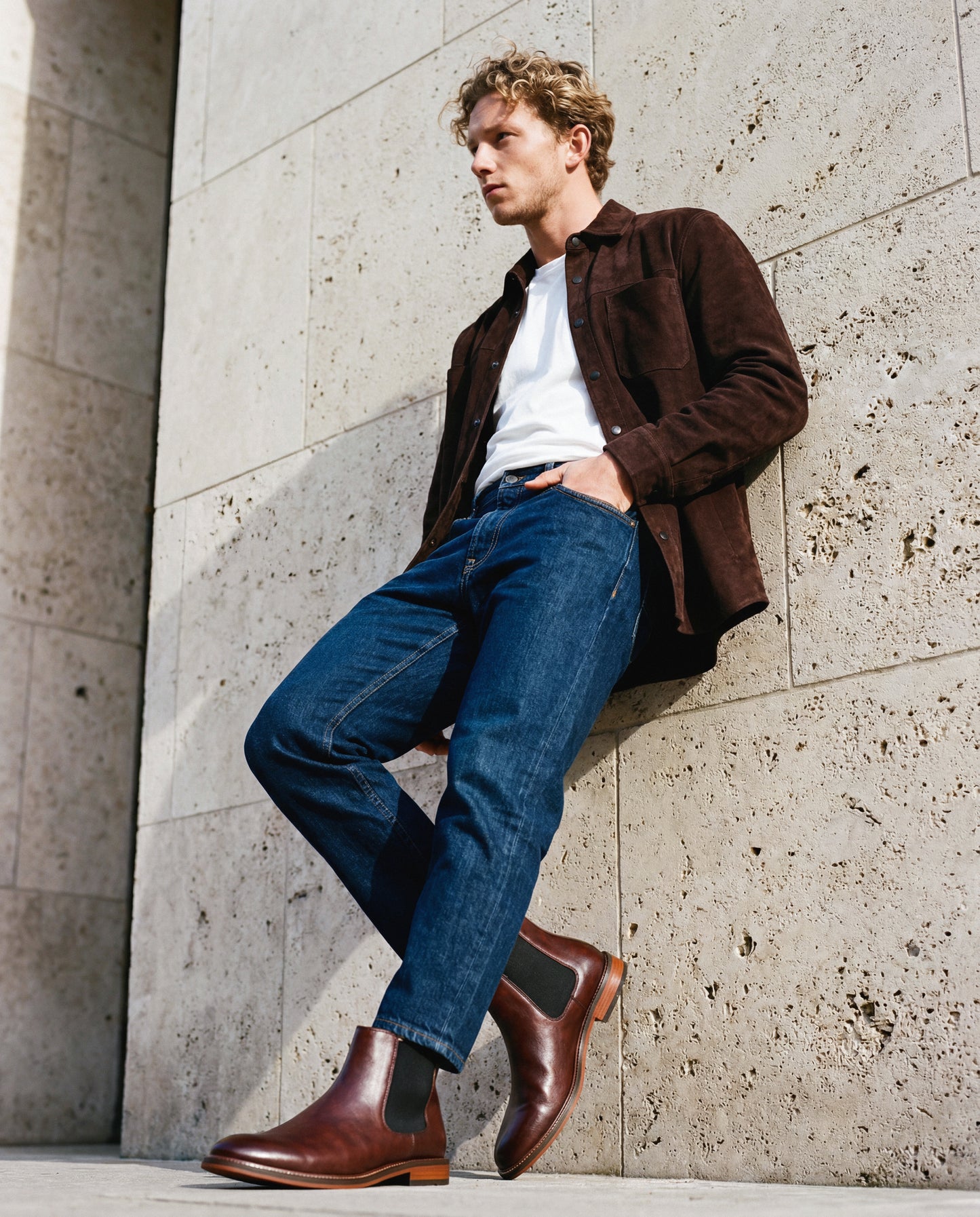 Man wearing a brown jacket, white shirt, blue jeans, and brown vegan boots from nae vegan shoes leaning against a stone wall.